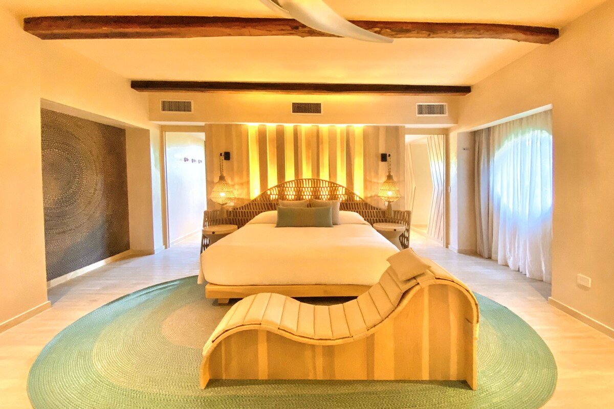 A spacious bedroom with a large bed, a curved bench, and a green rug.