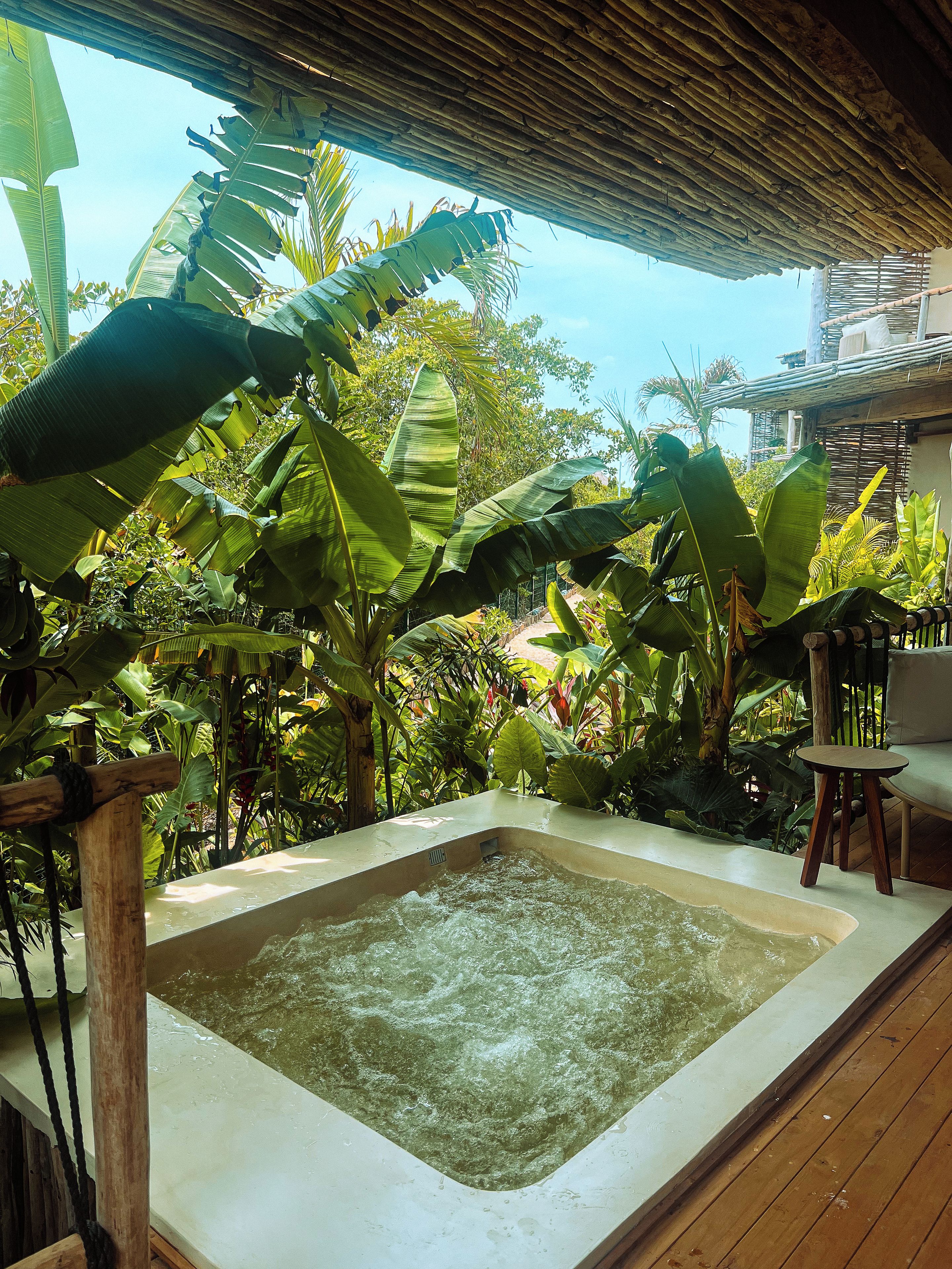 A hot tub surrounded by lush tropical foliage, with a wooden deck and a thatched roof overhead.