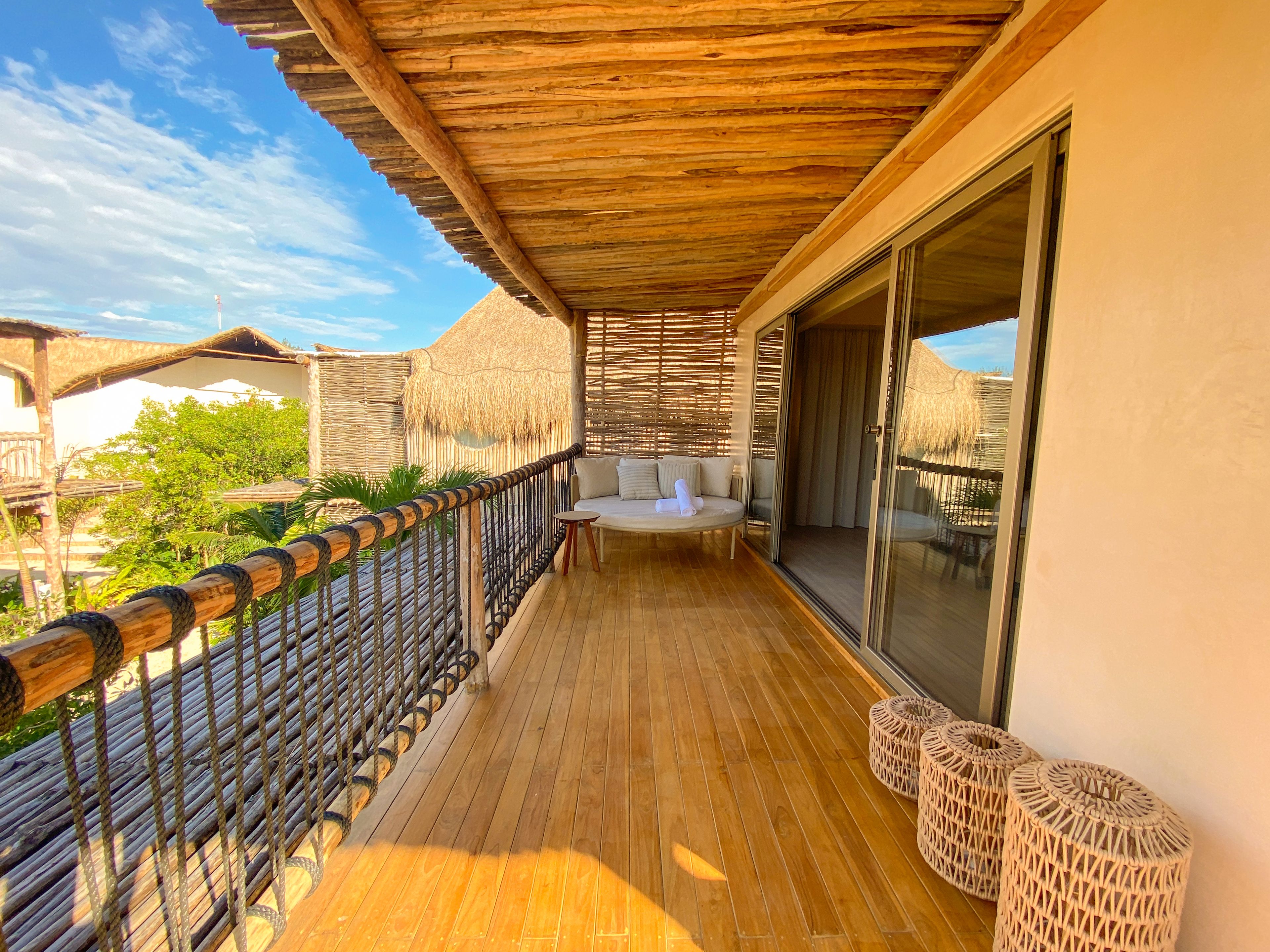 A wooden deck with a cushioned seating area, a glass door leading to a room, and a thatched-roof structure in the background.