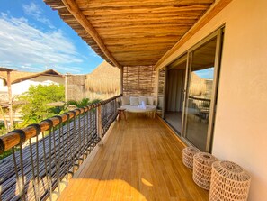 A wooden deck with a cushioned seating area, a glass door leading to a room, and a thatched-roof structure in the background. - Hotel Shibari Tulum (Tulum)
