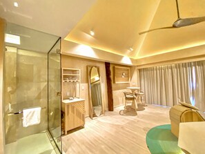 A modern hotel room with a glass shower enclosure, a vanity with a mirror, a seating area with a table and chairs, and a large window with curtains. - Hotel Shibari Tulum (Tulum)