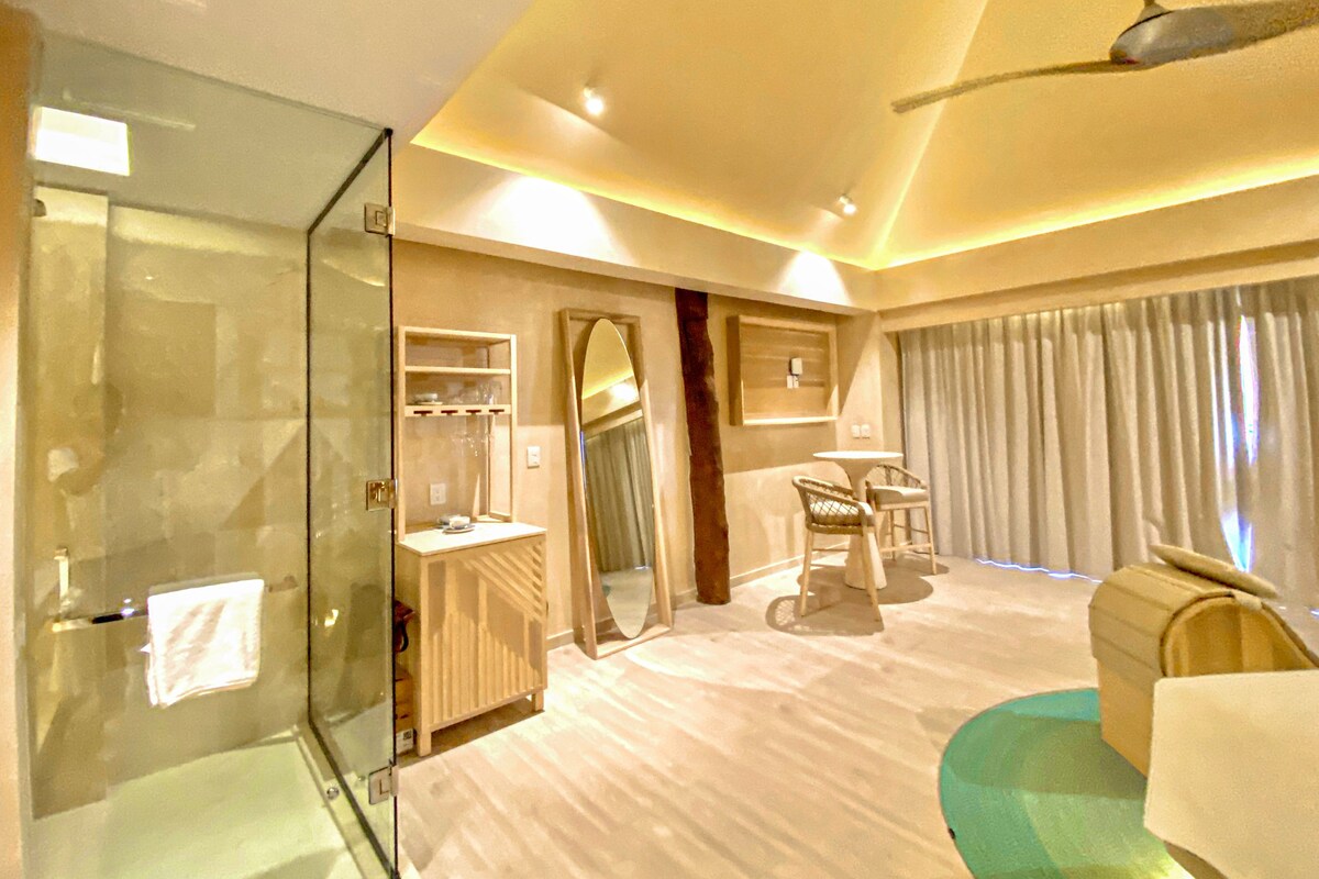 A modern hotel room with a glass shower enclosure, a vanity with a mirror, a seating area with a table and chairs, and a large window with curtains.