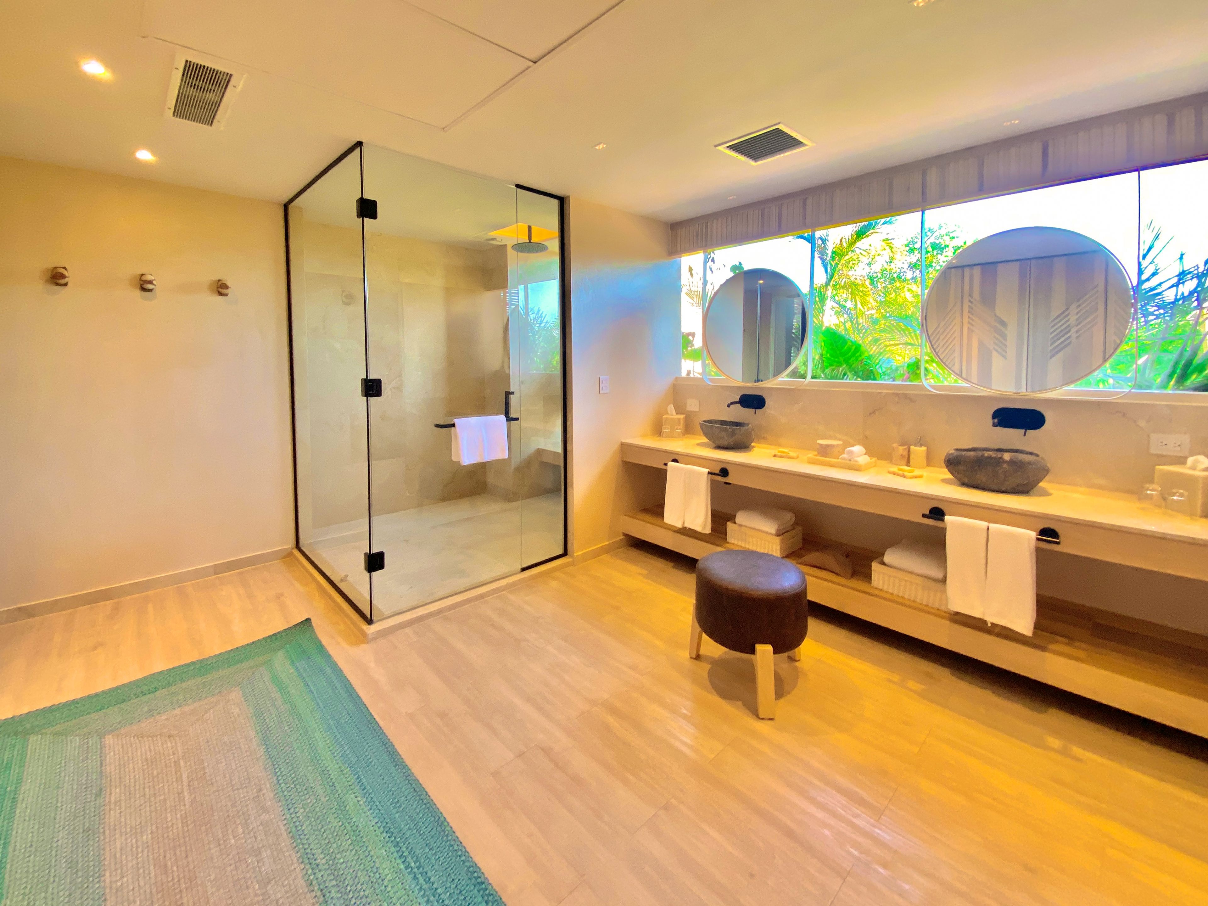 A modern bathroom with a glass shower enclosure, a vanity with a large mirror, and a view of greenery outside.