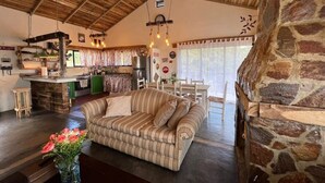 Een gezellige woonkamer met een gestreepte bank, een bijzettafeltje met een bloevas, een stenen openhaard en een keuken op de achtergrond.