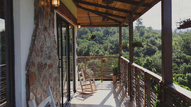 Een balkon met houten plafond, stenen muur en uitzicht op een bebost landschap.