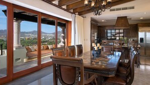 A spacious dining area with a large table and multiple chairs, a chandelier, and a view of the cityscape through large windows.