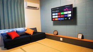 A modern living room with a dark blue sofa, orange and red cushions, a flat-screen TV mounted on a blue wall, a clock, and a Wi-Fi sign.