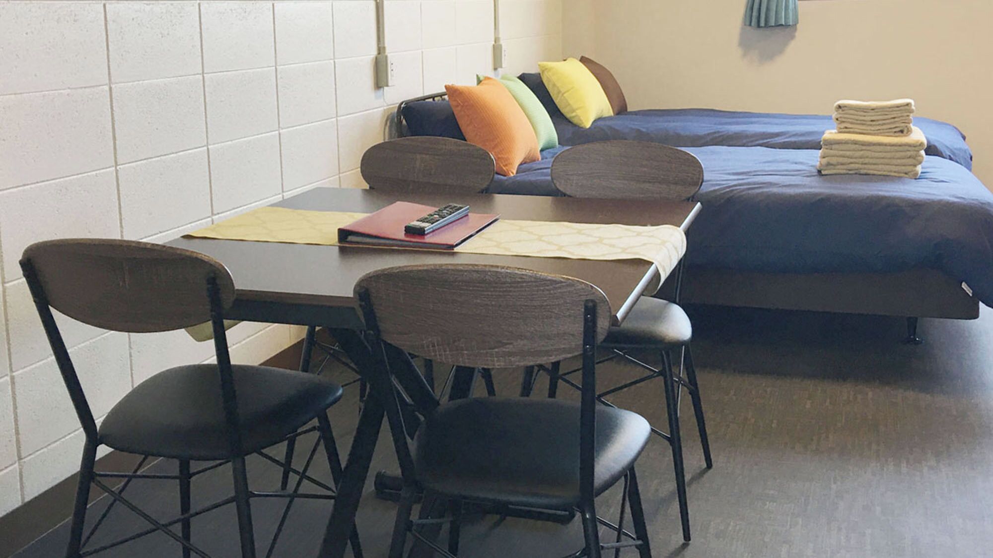 A room with a bed, two chairs, a table, and a small desk with a remote control and a book.