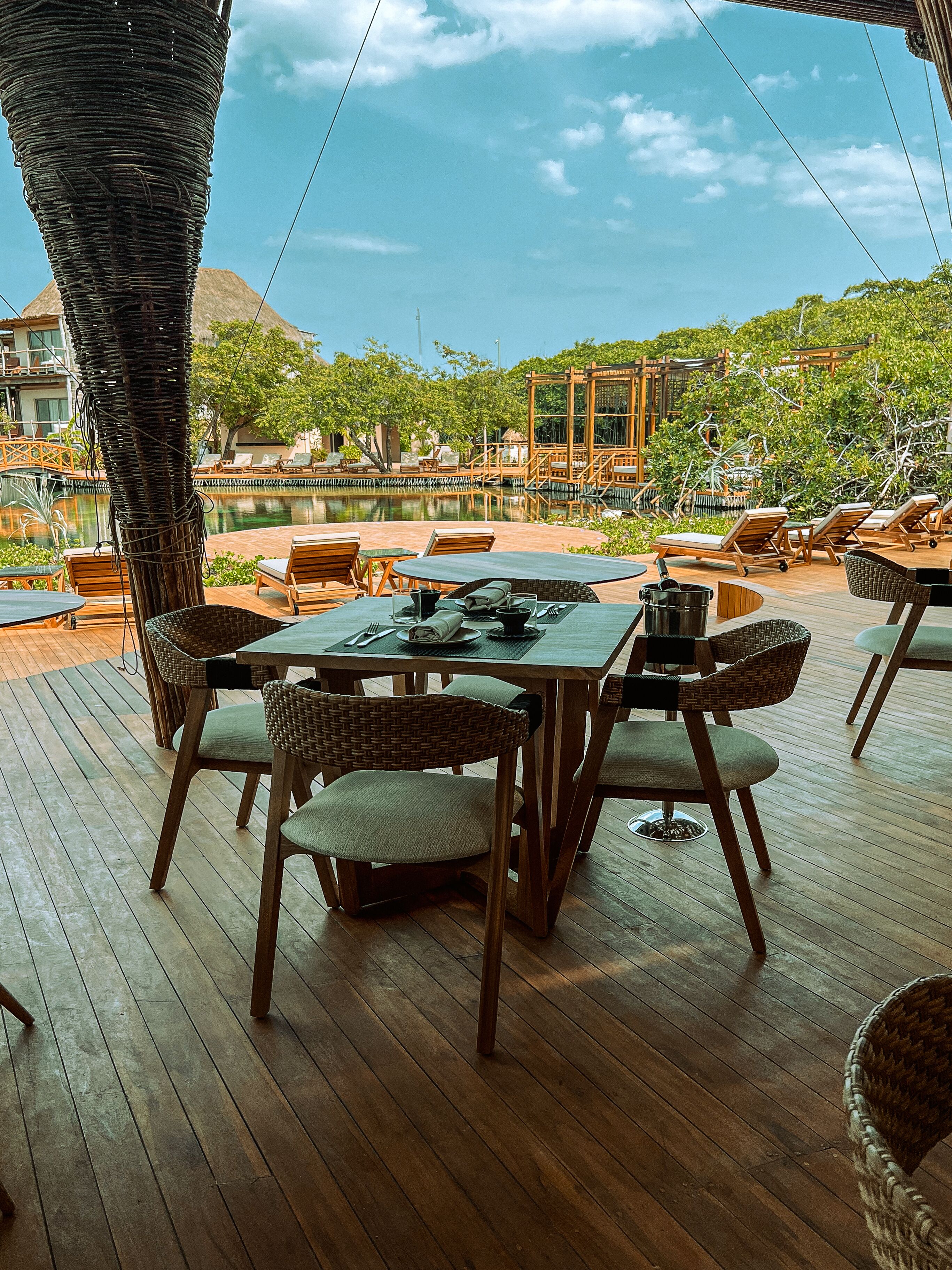 An outdoor dining area with wooden tables and chairs, a pool, and a view of a tropical landscape.