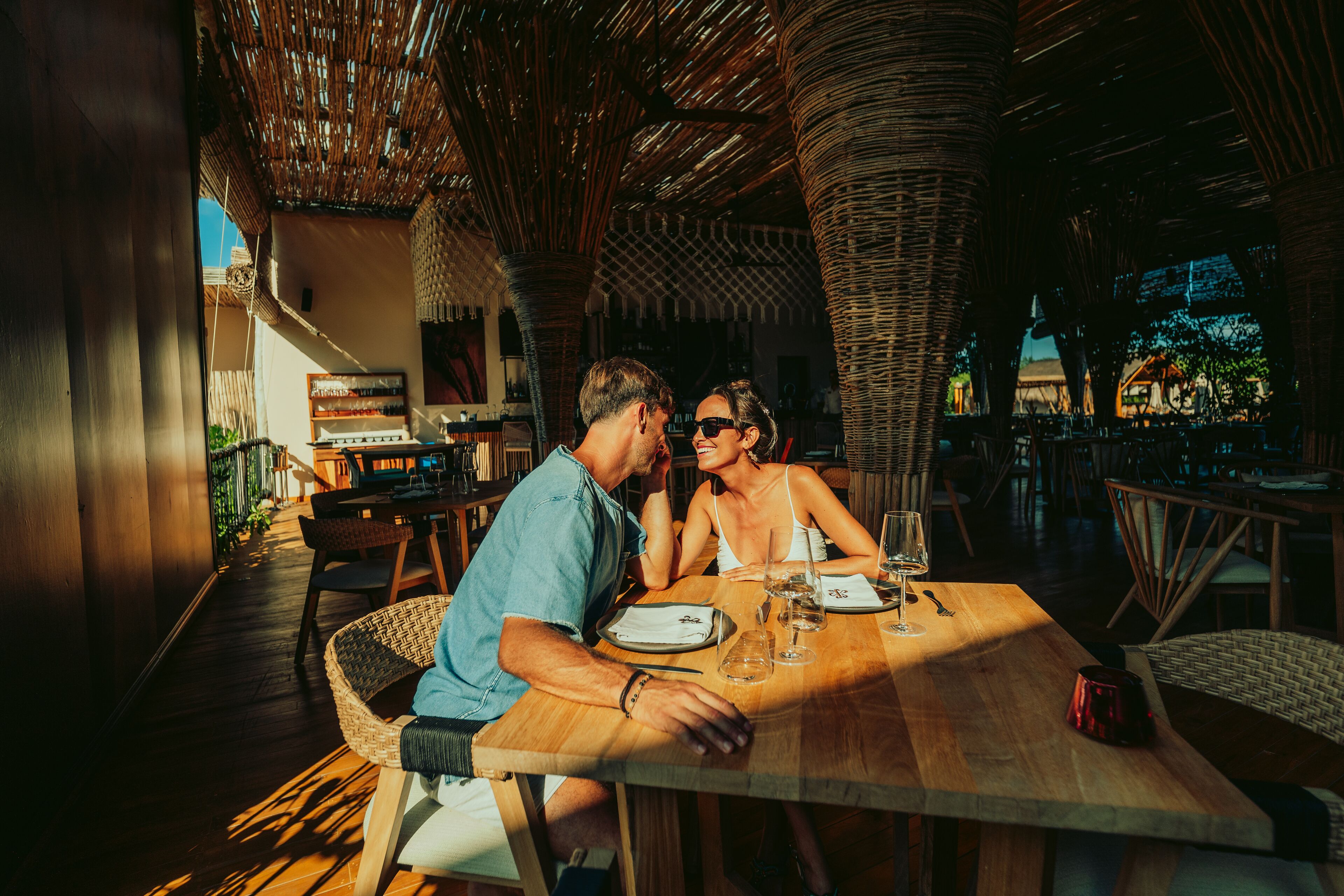 A couple dining at an outdoor restaurant with wicker furniture and a wooden table.