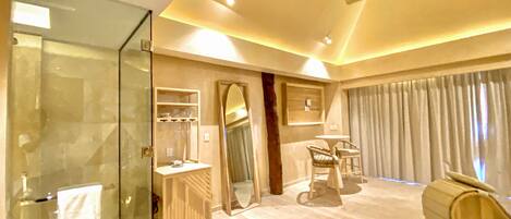 A modern hotel room with a glass shower enclosure, a vanity with a mirror, a seating area with a table and chairs, and a large window with curtains.