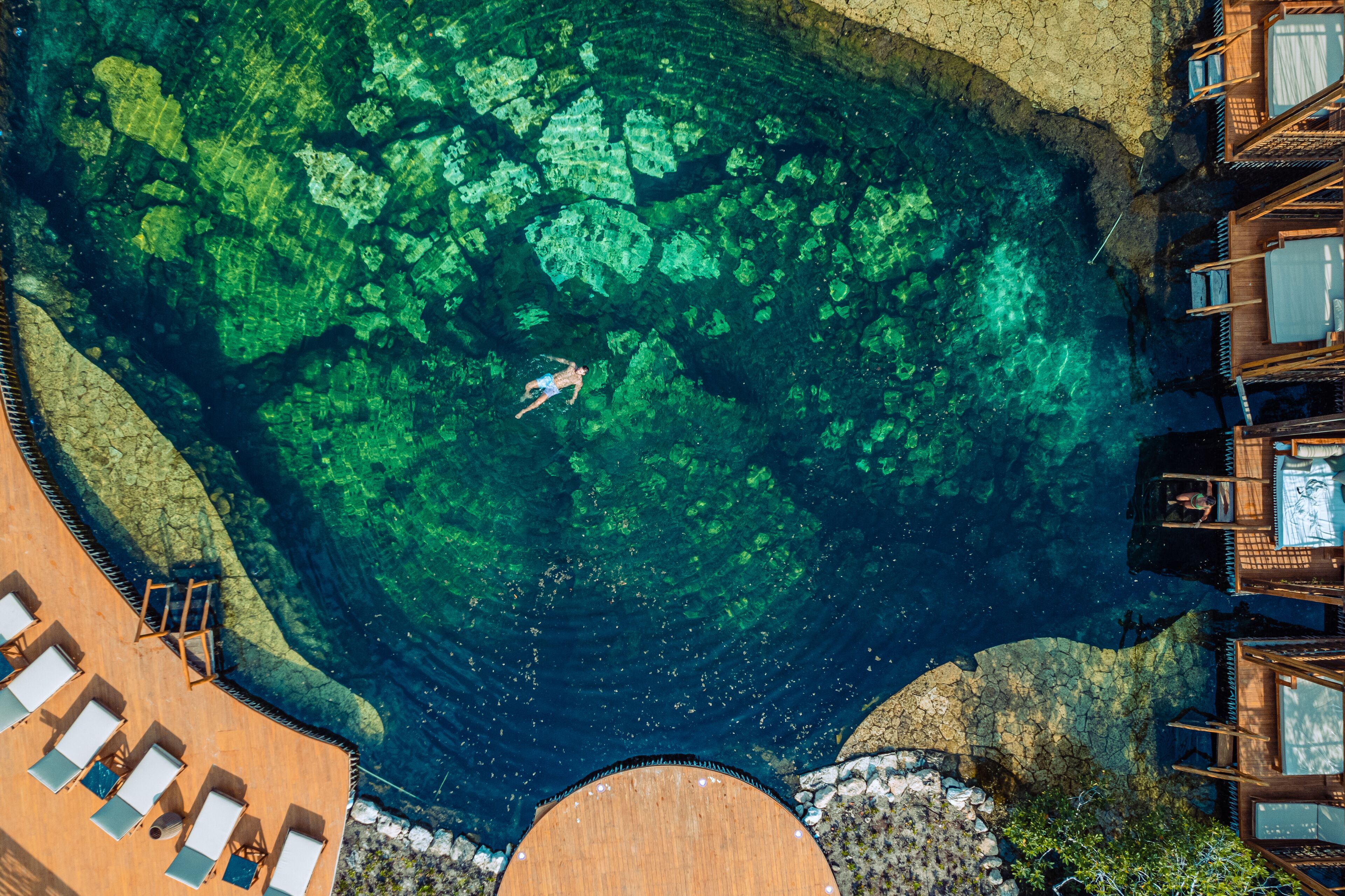 An aerial view of a resort with clear blue water, wooden deck, and lounge chairs.