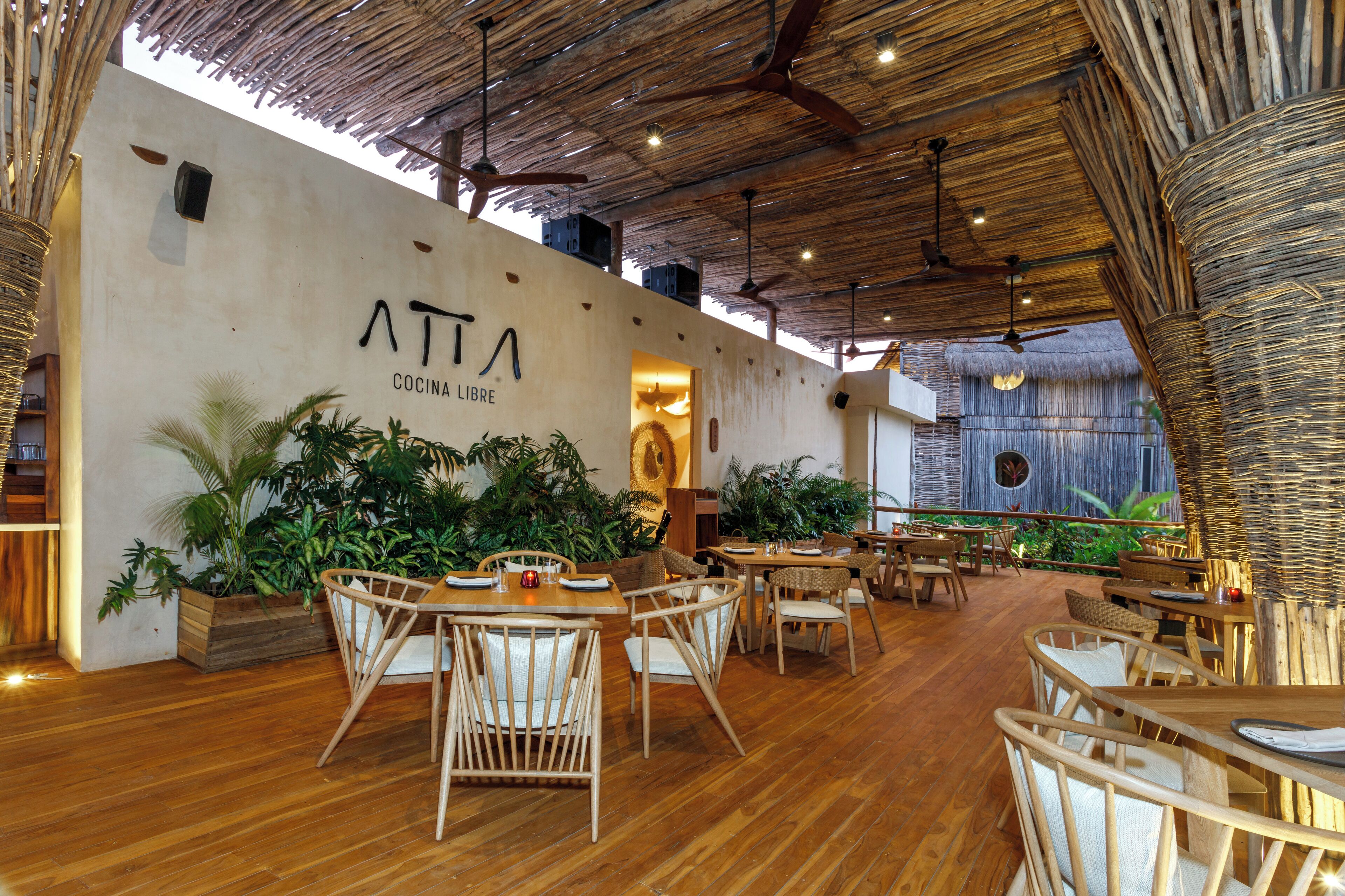A restaurant interior with wooden flooring, wicker furniture, and a thatched ceiling.