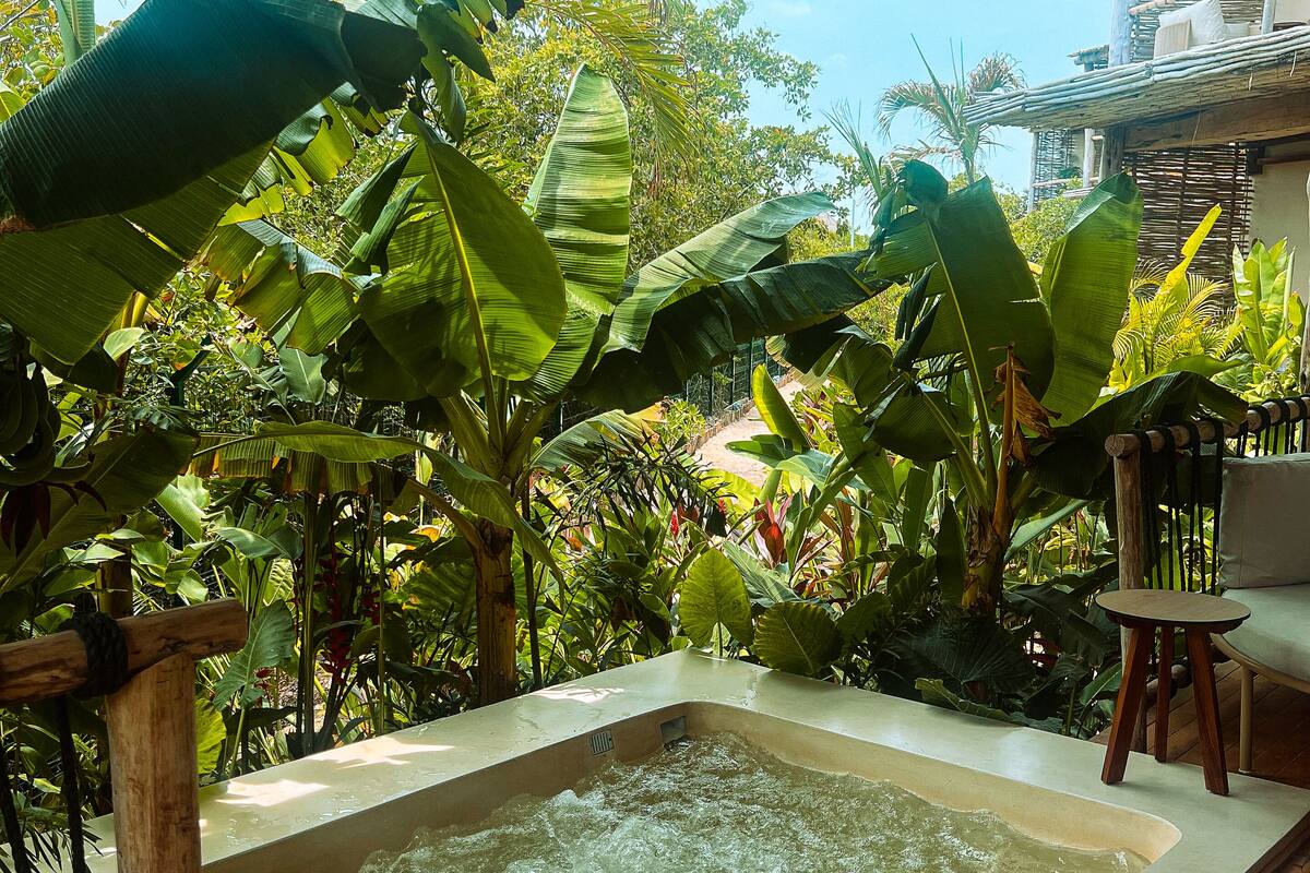 A hot tub surrounded by lush tropical plants, with a wooden deck and a thatched roof overhead.
