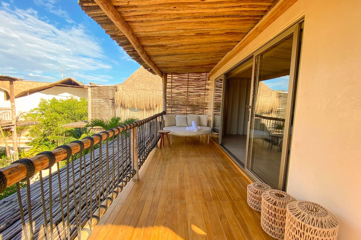 A wooden deck with a cushioned seating area, a glass door leading to a room, and a thatched-roof structure in the background.