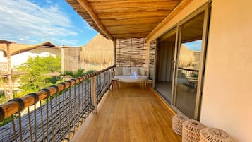 Una terraza de madera con zona de estar acolchada, un ventanal de vidrio que da a una habitación y una estructura con techo de paja al fondo.