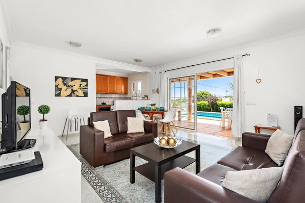 A modern living room with a brown leather sofa, a coffee table, and a TV. There is a kitchen in the background and a sliding glass door leading to a pool area.