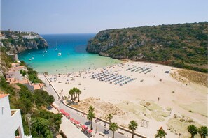 Une plage bordée de rangées de transats et de parasols, une route où sont garées des voitures, et une vue sur la mer et les falaises.