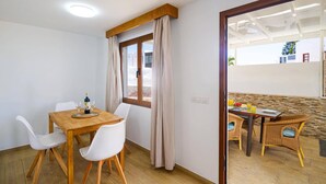 A dining area with a wooden table and white chairs, a window with curtains, and a view of a building outside. - Villa Fayna by Villa Plus (Puerto del Carmen)