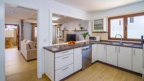 A modern kitchen with white cabinets, a stainless steel dishwasher, and a microwave. There is a dining area with a sofa and a table visible in the background. - Villa Fayna by Villa Plus (Puerto del Carmen)
