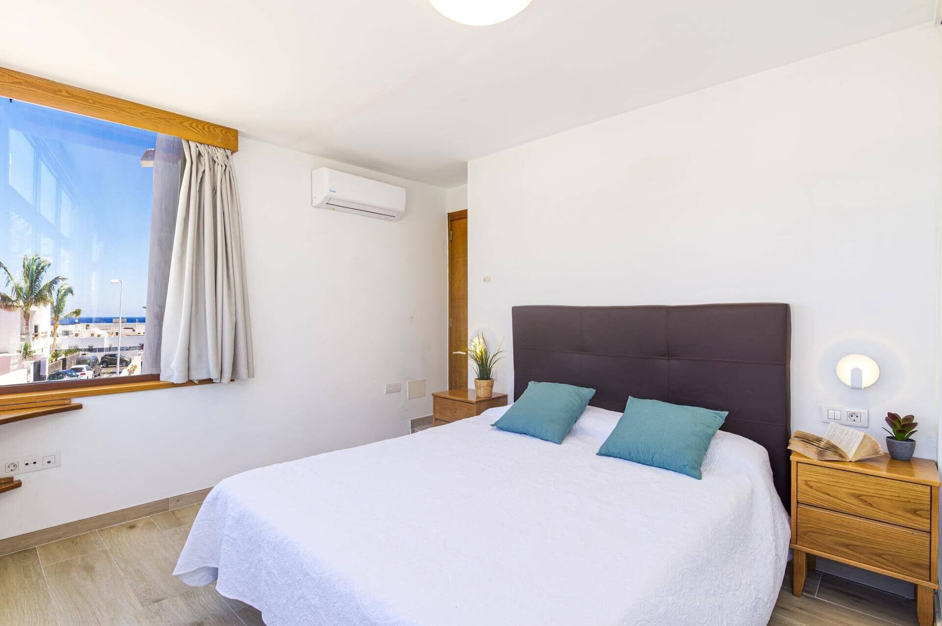 A bedroom with a bed, a nightstand, a bedside lamp, a window with curtains, and an air conditioning unit.