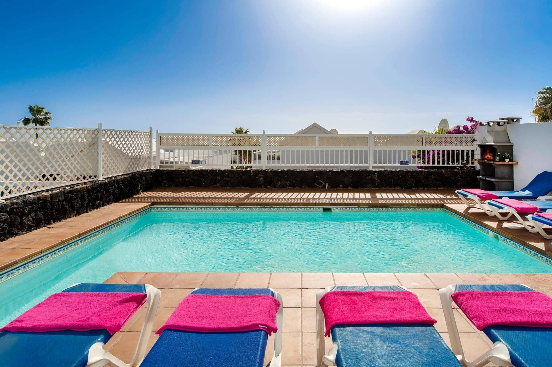 Une piscine entourée de chaises longues bleues et blanches, d’une clôture blanche et d’un mur en pierre.