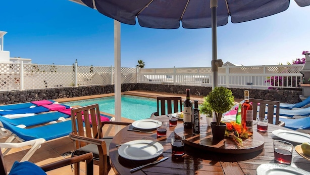 A poolside area with lounge chairs, a table set for dining, and a view of the pool and ocean. - Villa Siesta by Villa Plus (Puerto del Carmen)