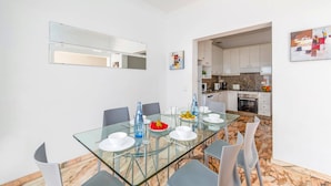 A modern dining area with a glass table and chairs, a kitchen in the background, and a white wall with a framed picture. - Villa Siesta by Villa Plus (Puerto del Carmen)