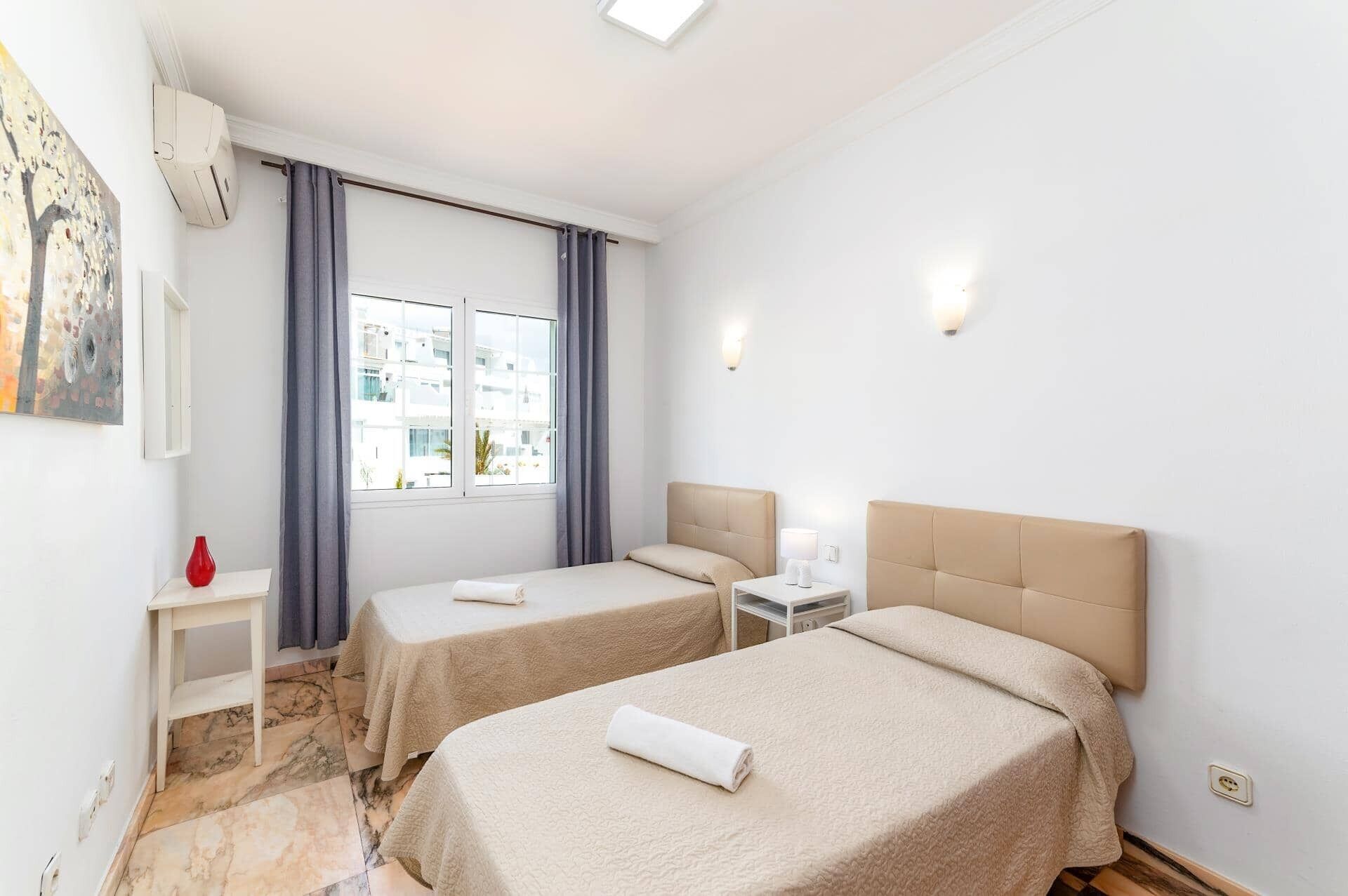 Une chambre d’hôtel avec deux lits, une petite table, un vase rouge et une fenêtre avec des rideaux.