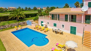 A pool area with multiple sun loungers and a dining table, surrounded by a pink building with green shutters. - Villa Daisy by Villa Plus (Lagos)