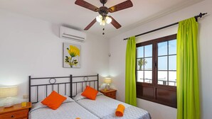 A bedroom with two beds, a ceiling fan, an air conditioning unit, a window with green curtains, and a painting on the wall. - Villa San Marcial by Villa Plus (Tías)