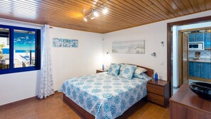 Een slaapkamer met een houten plafond, een bed met beddengoed met een patroon, een nachtkastje, een raam met uitzicht en een deur naar een keuken.