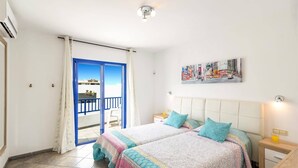 Een kamer met twee bedden, een balkon met een blauwe reling, een schilderij van een stadsgezicht en een betegelde vloer.