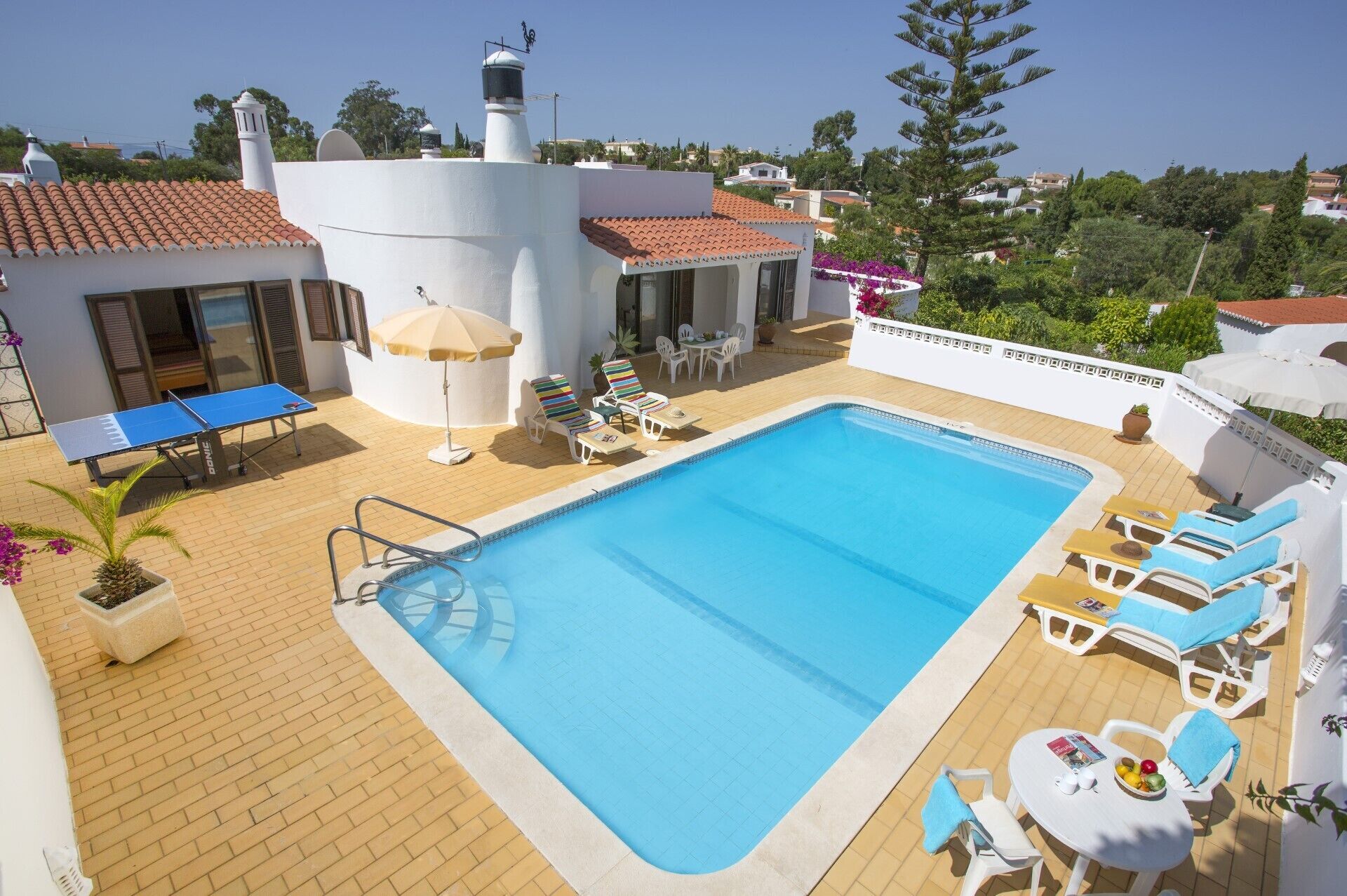 A swimming pool with lounge chairs and a ping pong table, surrounded by a white building with a tiled roof.