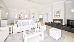 A modern living room with a white sofa, coffee table, and a fireplace. There is a kitchen area visible in the background. - Villa Citrinos by Villa Plus (Almansil)