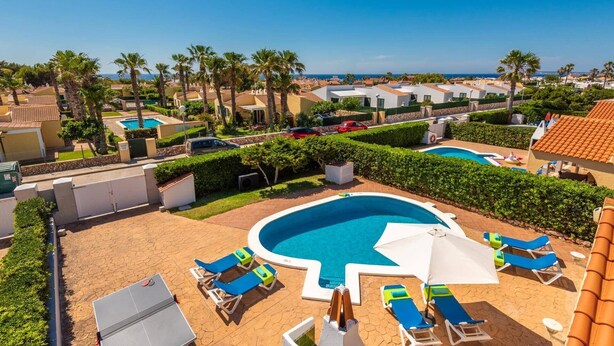 A resort with a central swimming pool, lounge chairs, and palm trees. - Villa Pluton by Villa Plus (Cap d'Artrutx)