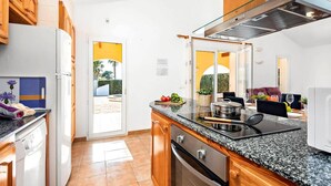 A modern kitchen with a built-in stove, oven, and microwave. There is a dining area with a table and chairs, and a view of the outdoors through a glass door. - Villa Pluton by Villa Plus (Cap d'Artrutx)