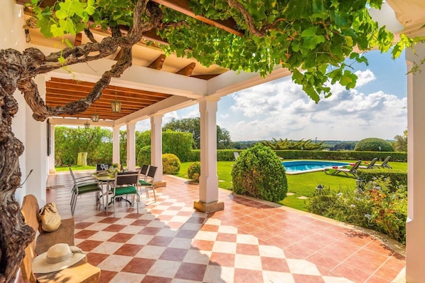 Un porche cubierto con piso a cuadros, mobiliario exterior y vistas a una piscina y jardín.