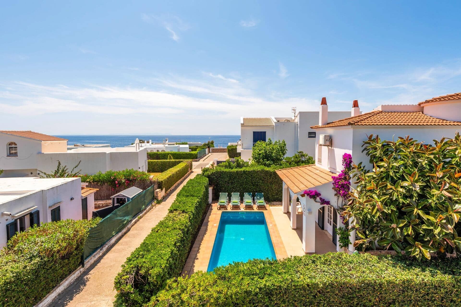 A pool area with a clear view of the sea, surrounded by white buildings with terracotta roofs, and a well-maintained hedge along the pool's edge.