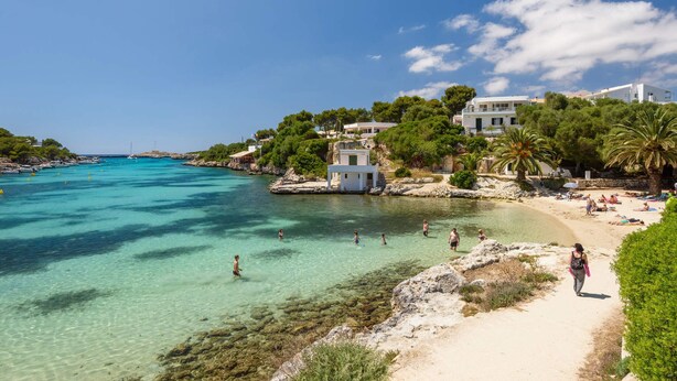 Eine Küstenlandschaft mit klarem türkisblauem Wasser, einem Sandstrand und Gebäuden mit Balkonen, die auf das Meer blicken.
