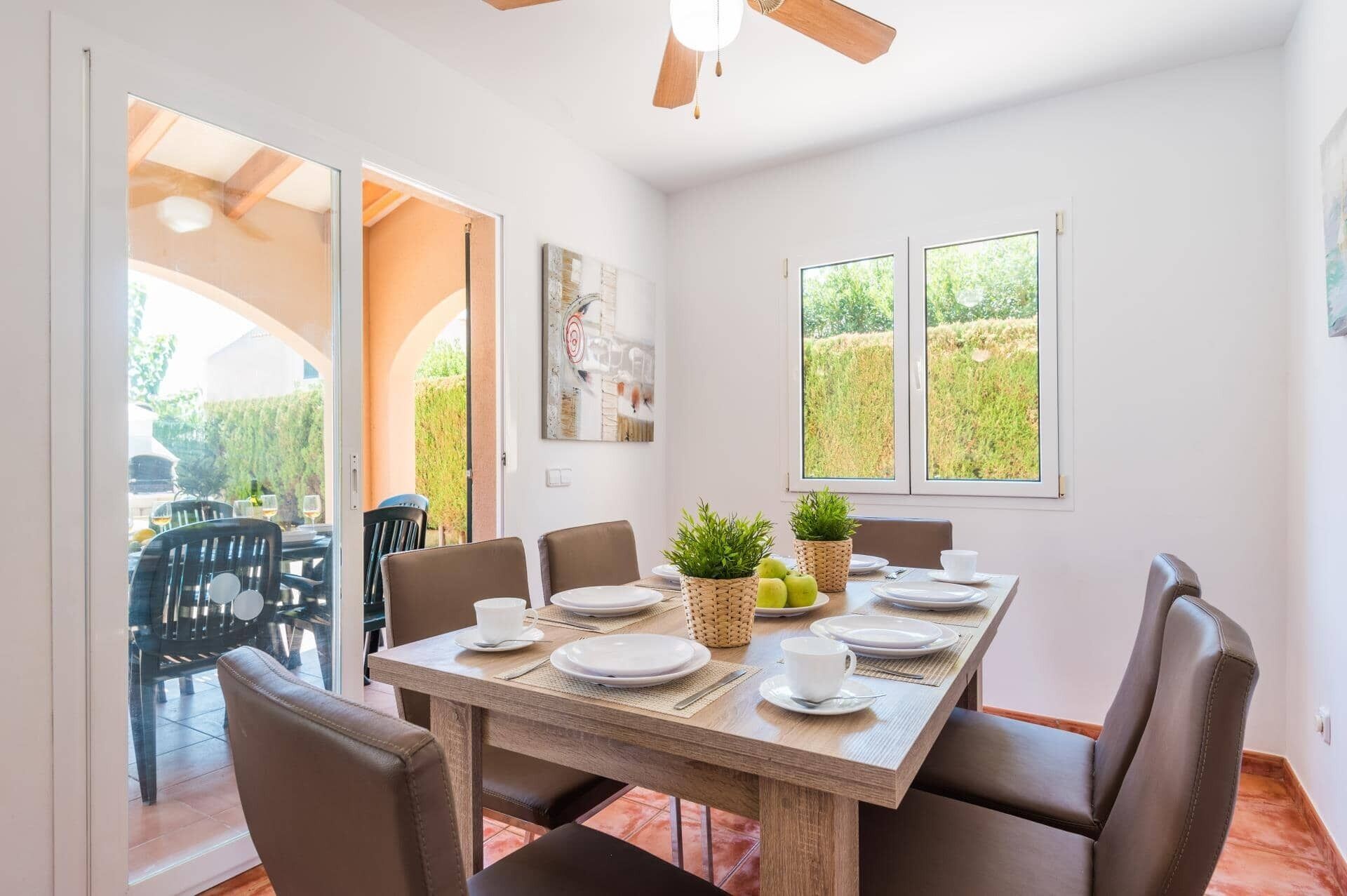 Un comedor con una mesa de madera dispuesta para cuatro personas, rodeada de sillas de cuero. Hay dos plantas en macetas sobre la mesa, y un cuadro en la pared.
