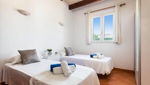 A hotel room with two beds, a window with curtains, a nightstand with a vase and mugs, and a tiled floor.