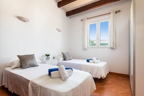 Une chambre d’hôtel avec deux lits, une fenêtre avec des rideaux, une table de chevet avec un vase et des tasses, et un sol carrelé.