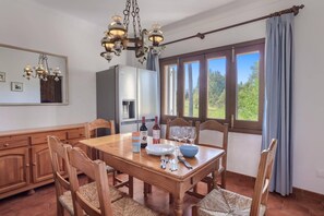 A dining area with a wooden table, chairs, a chandelier, and a view of greenery through large windows.