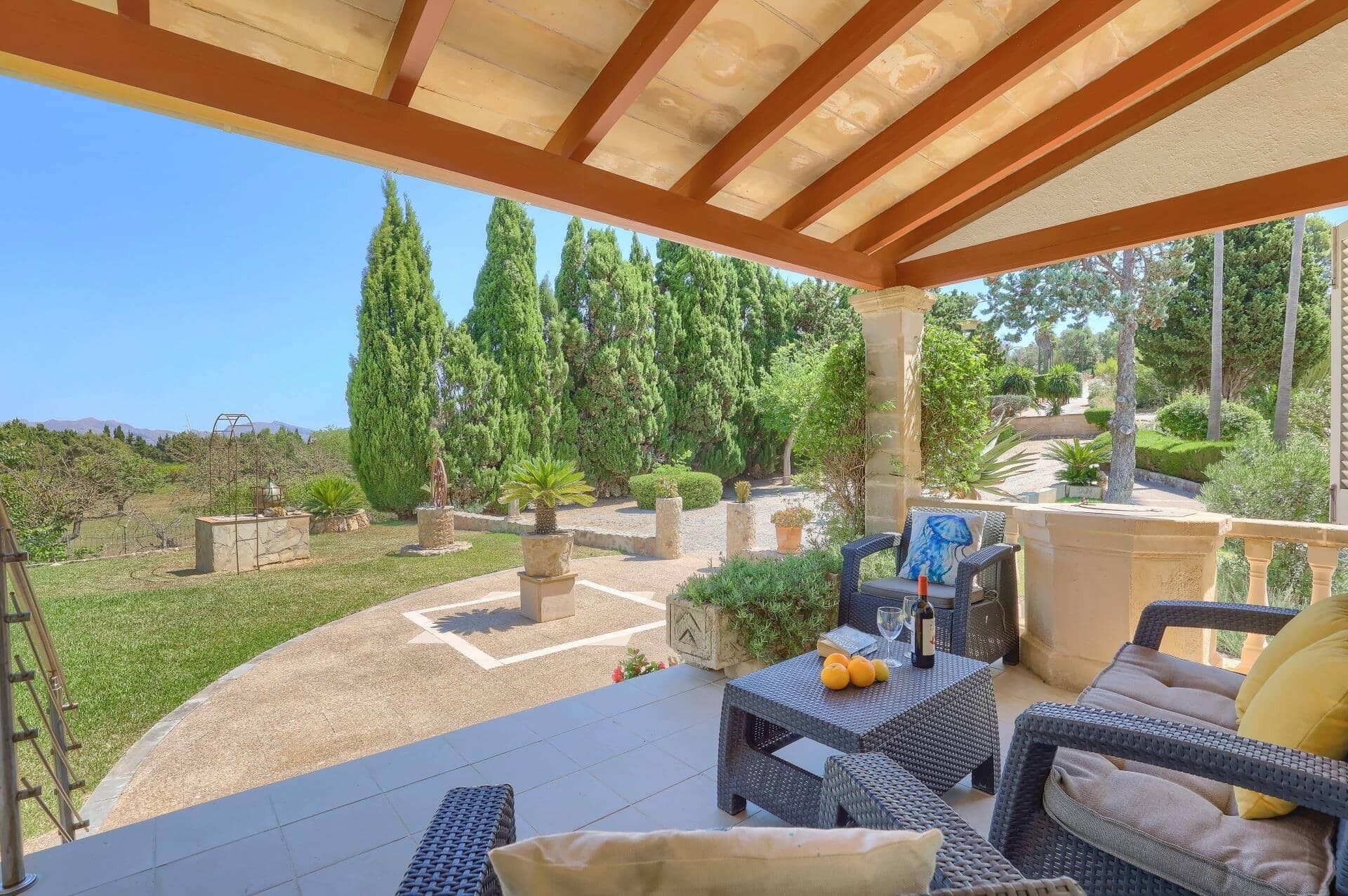 Un patio al aire libre con mobiliario de mimbre, una mesa con frutas y una botella, y vistas a un jardín ajardinado con árboles y un cielo despejado.
