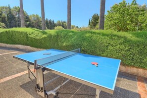 A table tennis table with a net, placed outdoors with a hedge and trees in the background.