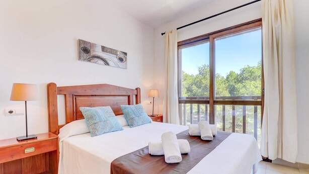A well-lit hotel room with a large bed, two bedside tables, a wooden headboard, and a large window with a view of trees.