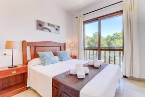 A well-lit hotel room with a large bed, two bedside tables, a wooden headboard, and a large window with a view of trees. - Villa Mar Egos by Villa Plus (Cala d'Or)