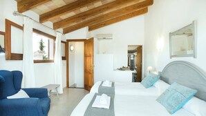 A bedroom with two beds, a wooden ceiling, a bathroom visible through an open door, and a blue armchair. - Villa Mari Pollensa by Villa Plus (Mallorca)