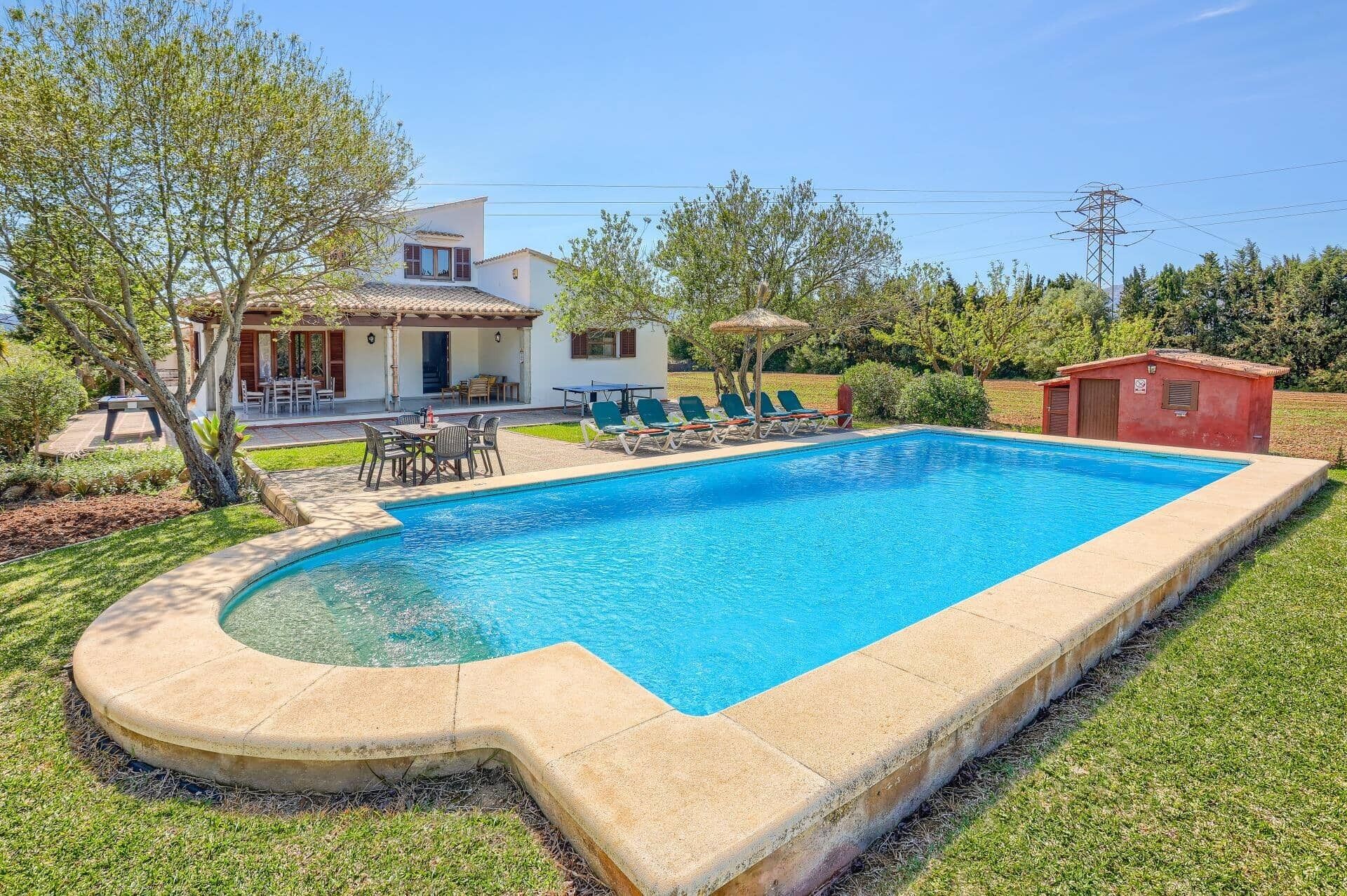 A swimming pool with a sunbathing area, a dining table, and a small red building.